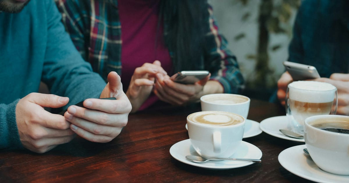 Three friends holding smartphones over cups of coffee on a wooden café table.