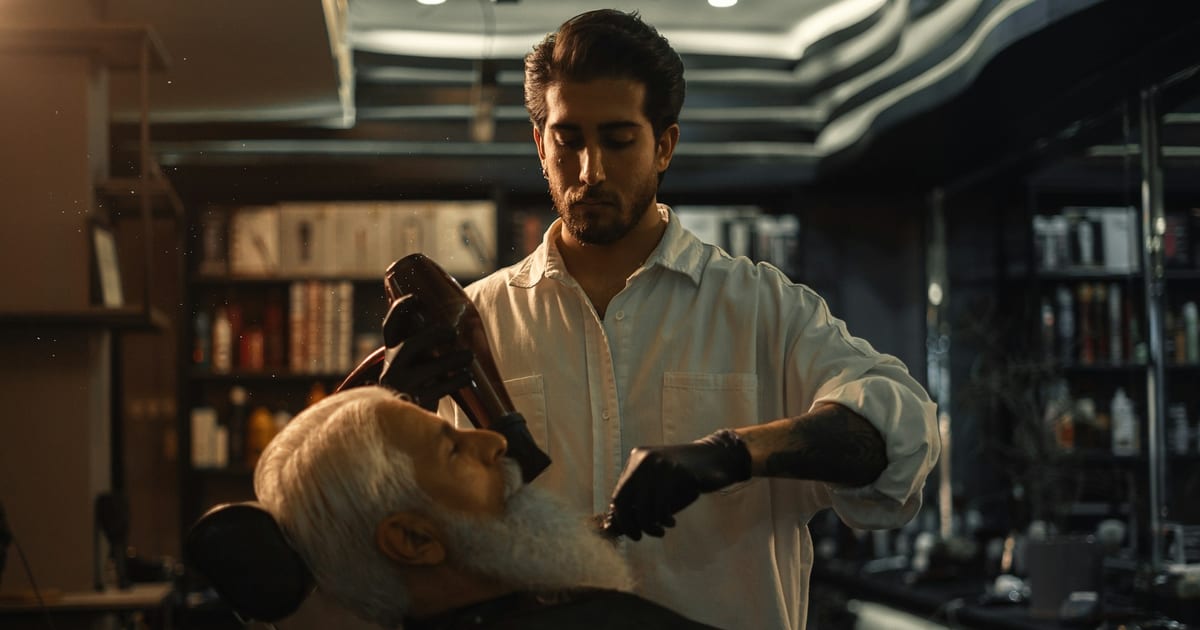 A barber trimming an older client's beard in a barbershop chair.