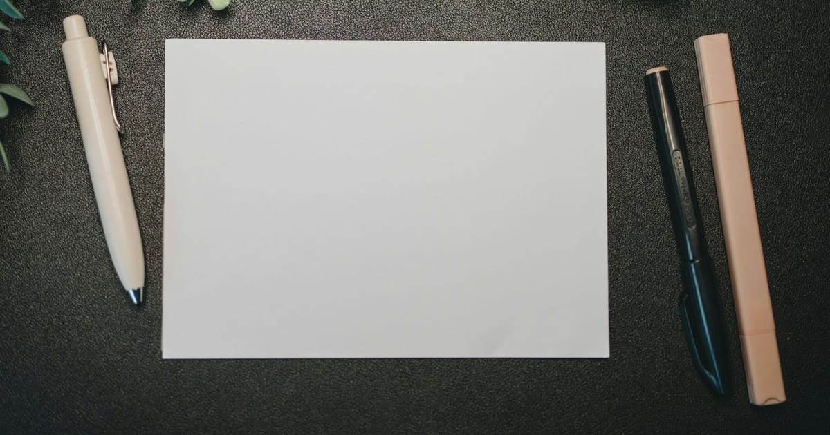 A blank white card on a dark desk between a pen, a marker, and a highlighter.