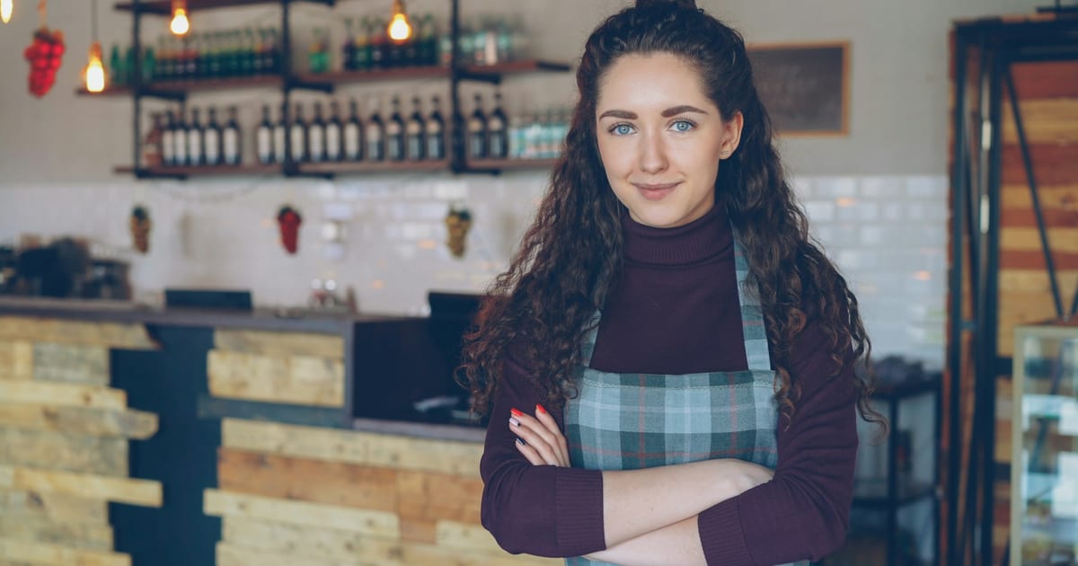 A cafe owner in an apron standing with her arms folded in front of her counter.