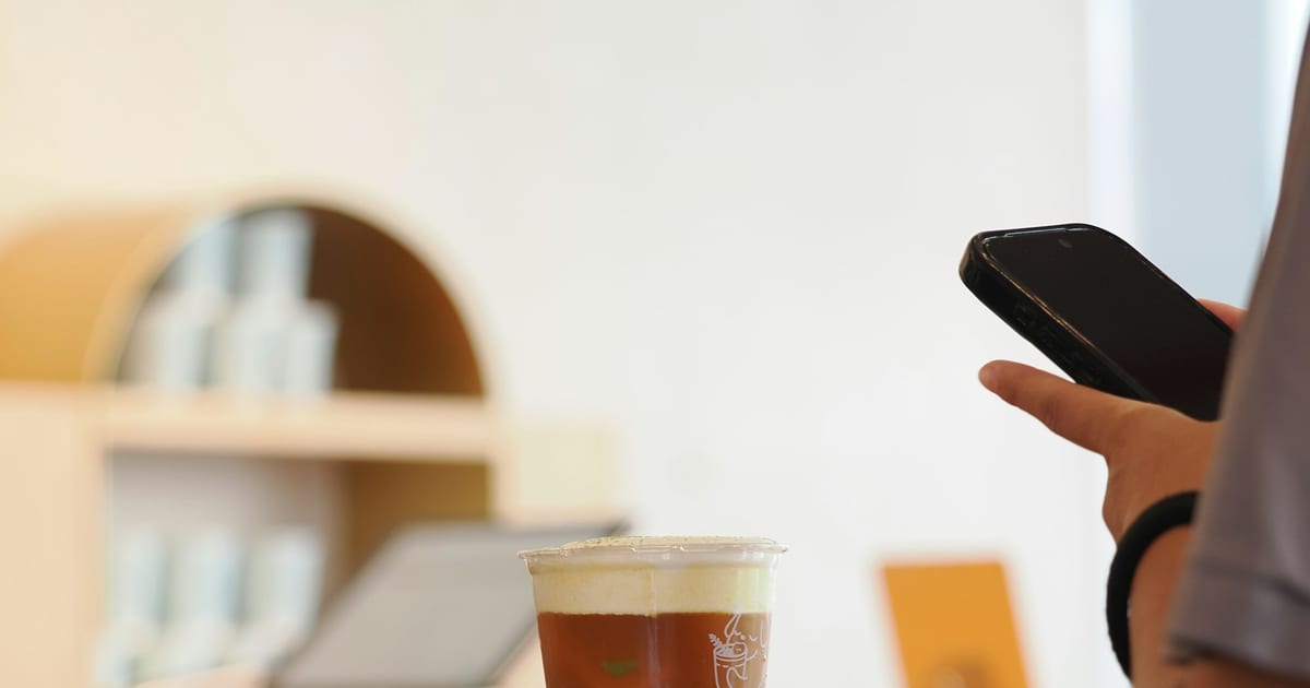 A customer holding a phone at a café counter next to an iced coffee.