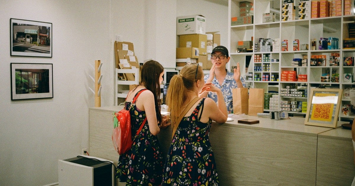 Two new customers chatting with a shopkeeper across the counter of a small shop.