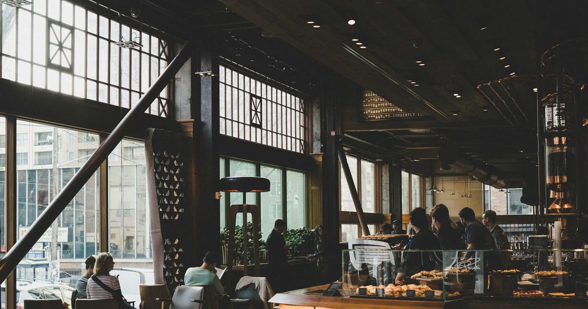 The inside of a large cafe with tall windows, a few customers at the counter and tables.
