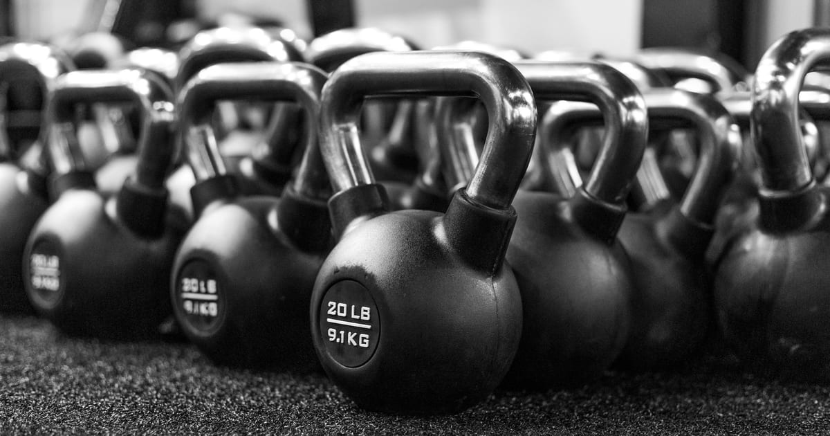 A row of black kettlebells lined up on the rubber floor of a gym, in black and white.
