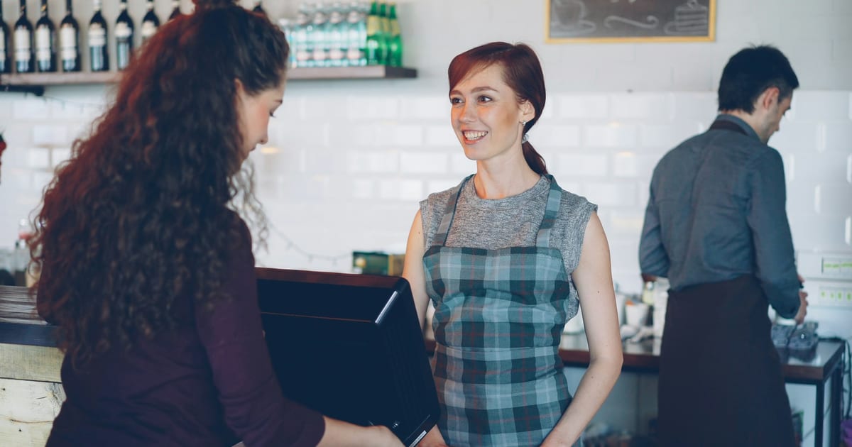 A smiling member of staff serves a customer at the counter of a small cafe while a colleague works behind her.