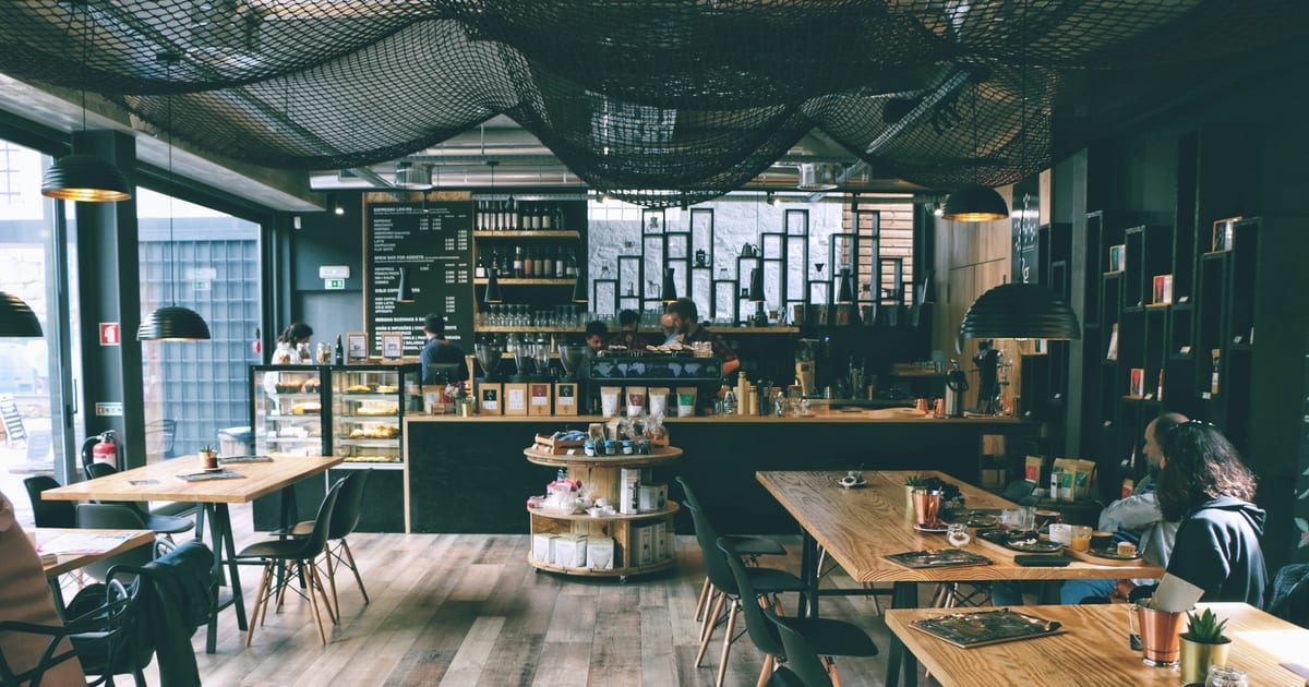 The inside of an independent coffee shop, with wooden tables, a long counter, a chalk menu board, and staff behind the bar.