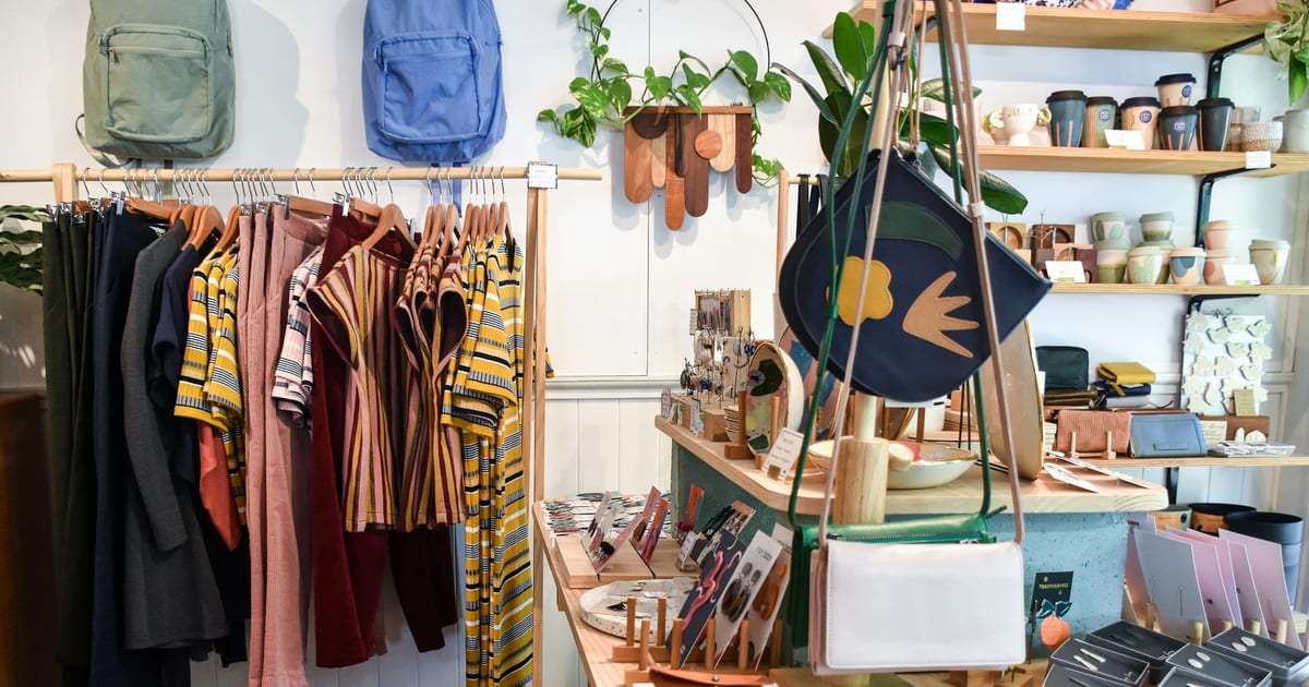 An independent shop with clothes on a wooden rail, bags on the wall, and ceramics on the shelves behind.