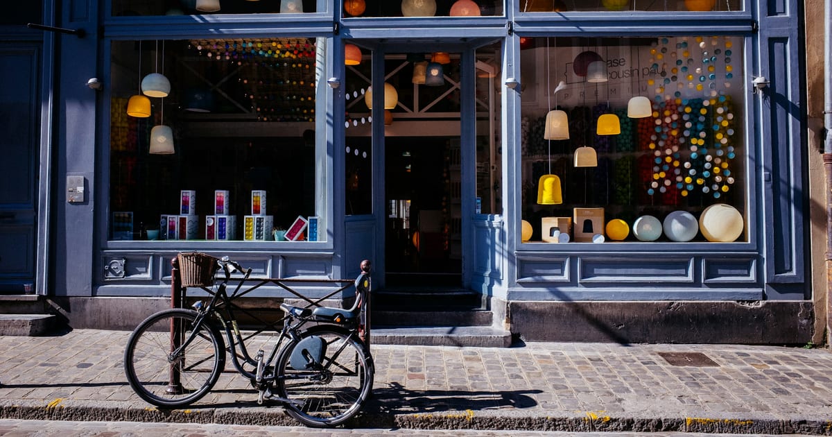 A bicycle parked on a cobblestone sidewalk outside the window of an independent lamp shop.