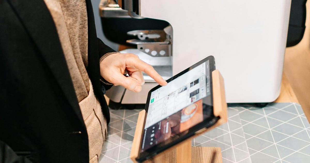 A hand tapping a till on a tablet in a wooden stand, beside a coffee machine.