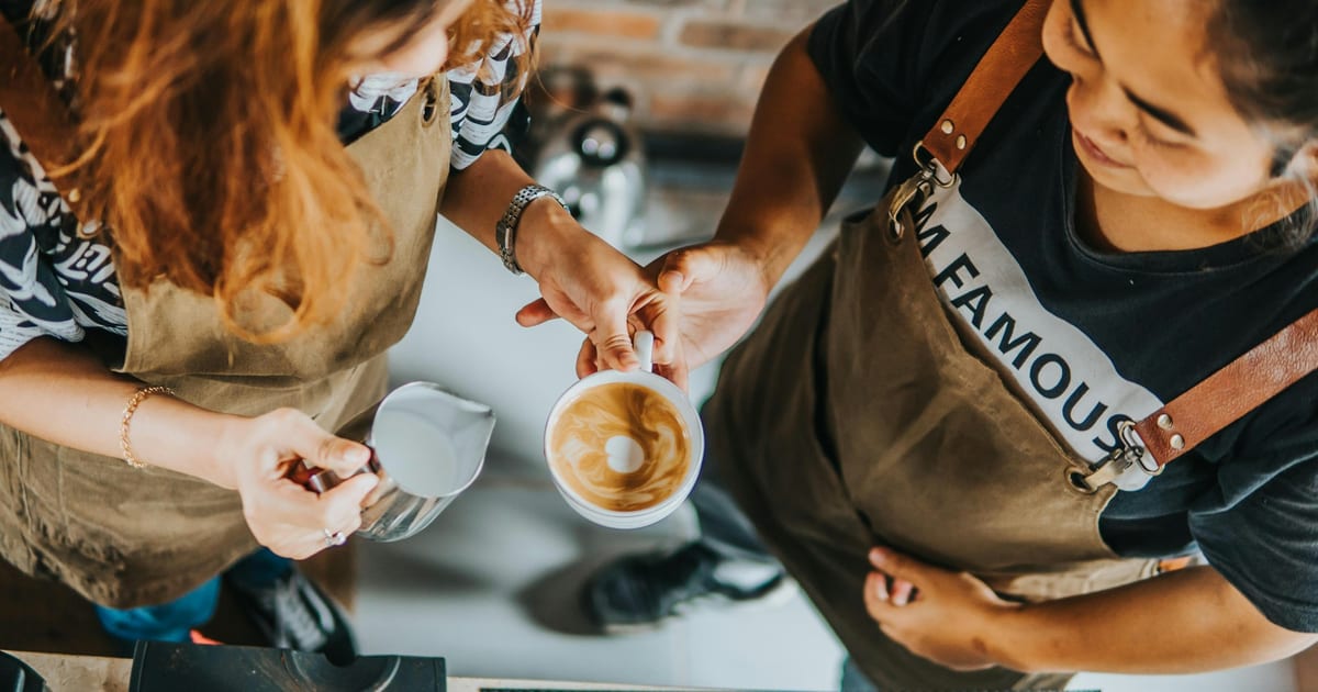 Two baristas at a coffee shop counter, one holding a finished latte.
