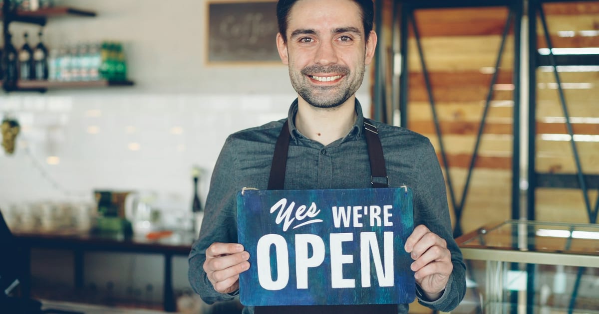 A smiling café owner holding up a "We're open" sign inside his coffee shop.