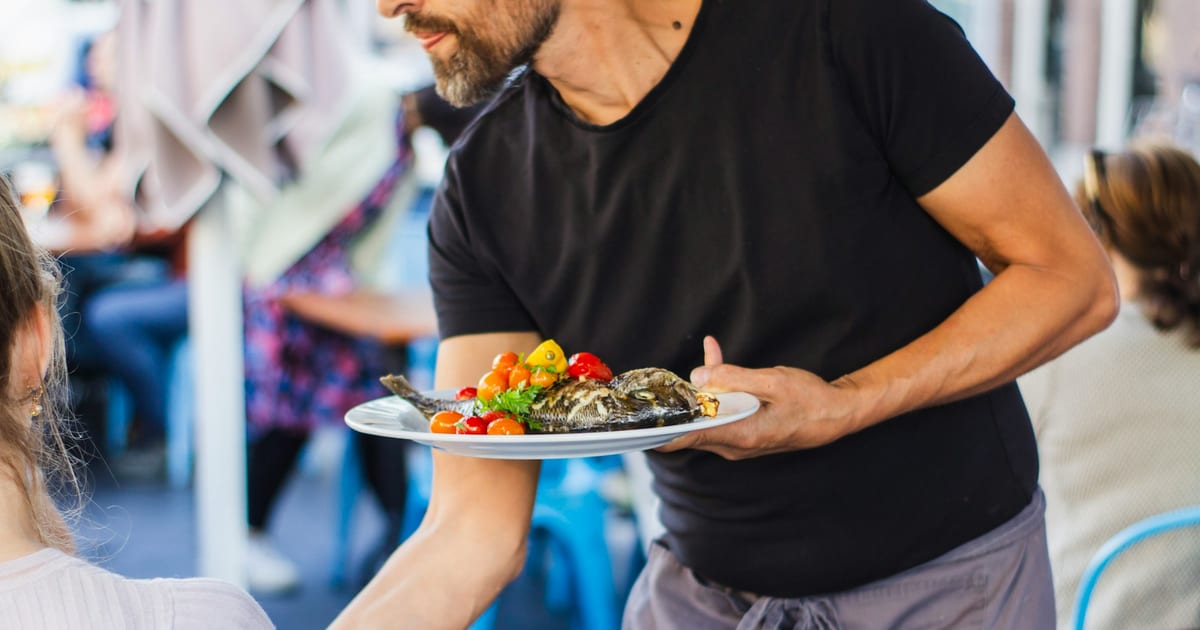 A server carrying a plate of grilled fish and vegetables to a table in a busy restaurant.