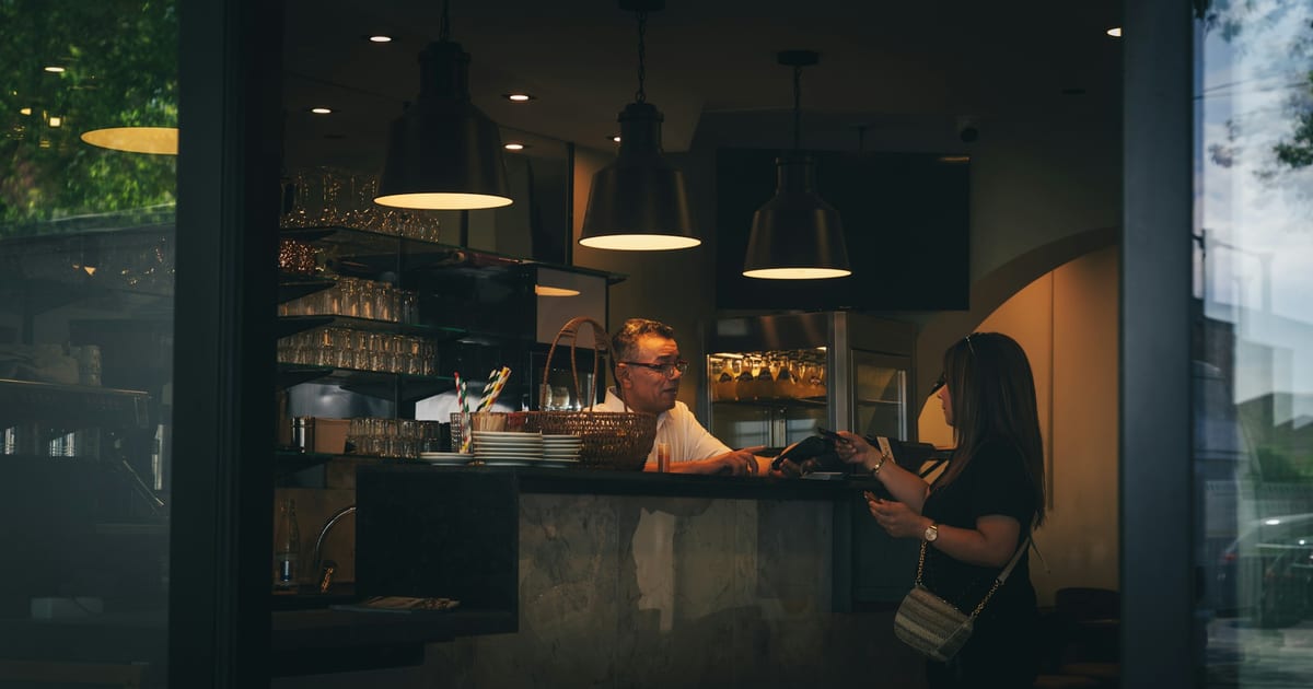A customer paying at the counter of a small café in the evening, seen through the front window.